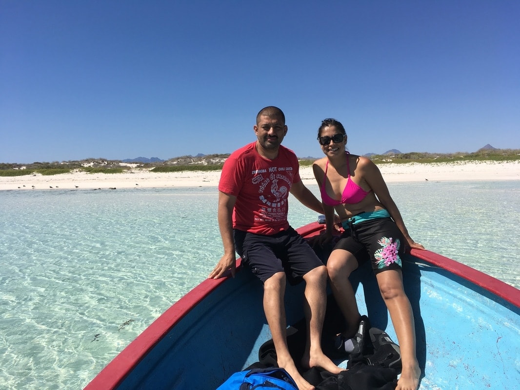 Couple sitting on the bow of a panga in impossibly clear crystal water with a white sand beach and sierra mountains behind near Loreto