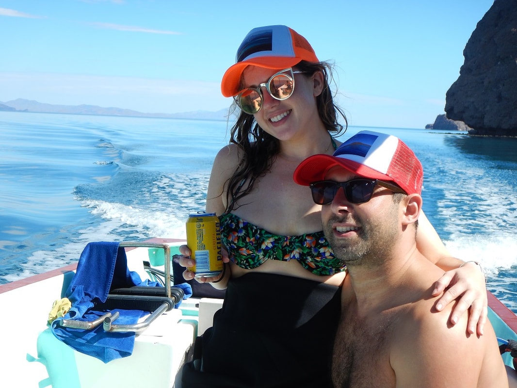 Happy couple in colorful trucker hats relaxing on a panga with turquoise gulf water and island cliffs behind on a Loreto tour