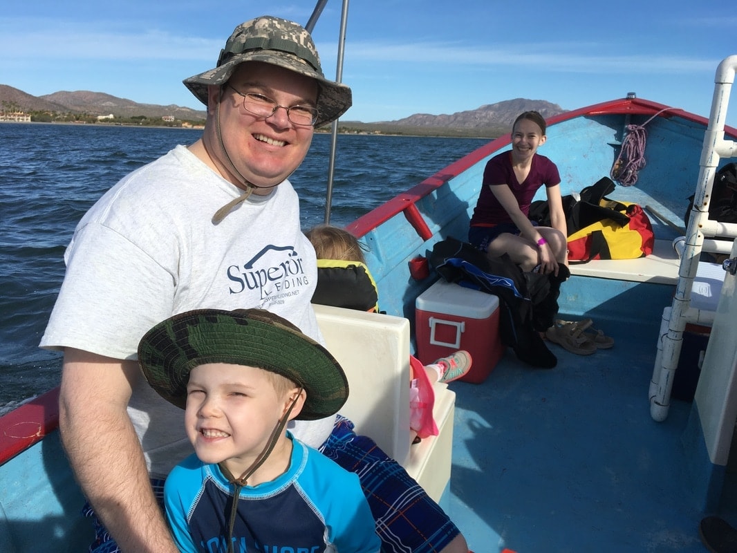 Dad and two young kids in sun hats smiling on a blue panga with sierra mountains and Loreto coastline in the background