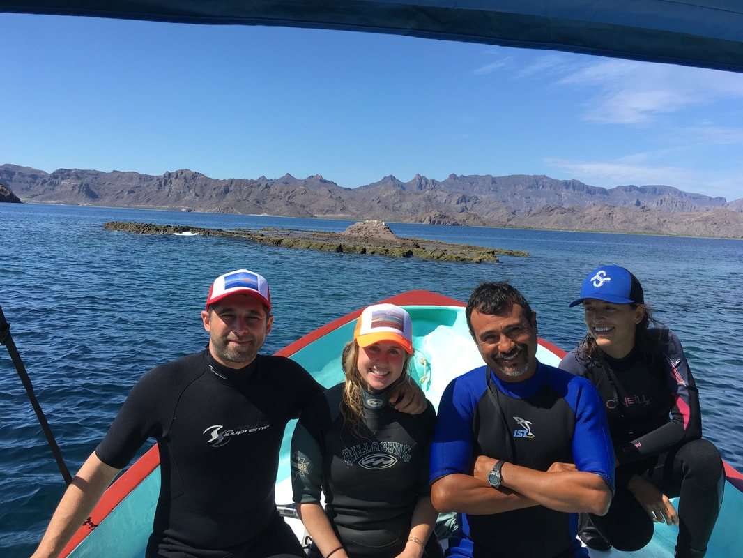 Four people in wetsuits smiling on a turquoise panga with a volcanic rock outcrop and sierra mountains behind them on a Loreto island tour