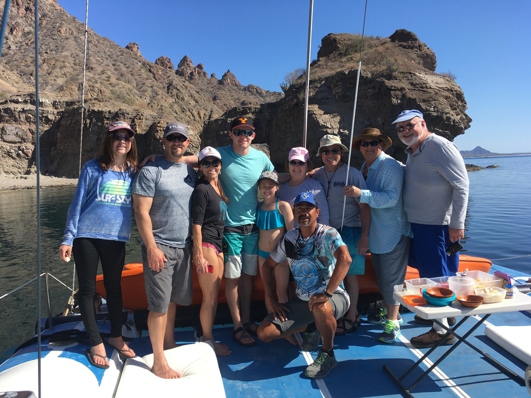 Group of nine guests and guide posing on a sailboat deck with dramatic rocky sierra cliffs rising behind them on a Loreto island tour