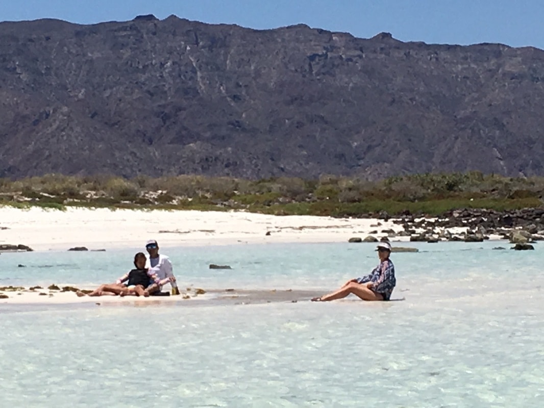 Three guests sitting in crystal-clear shallow water on a white sand island beach near Loreto