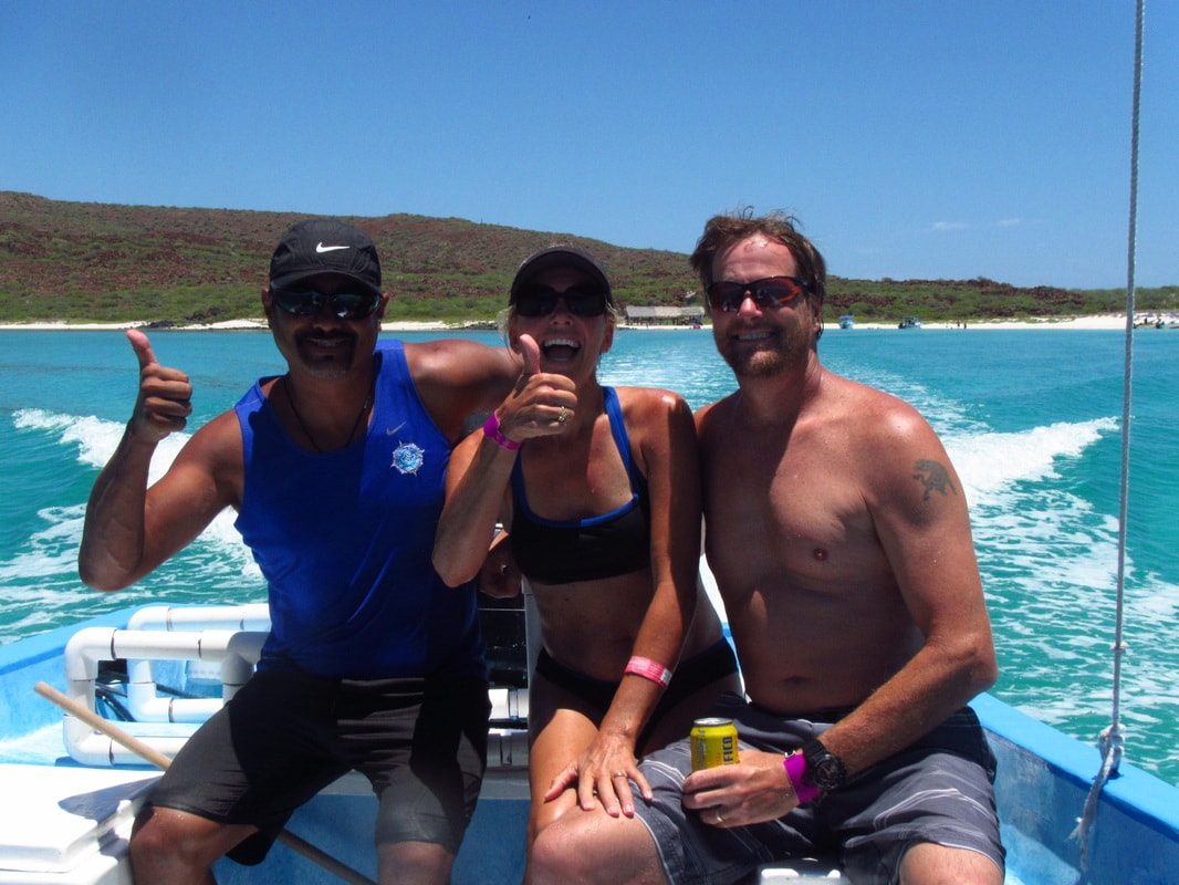 Guide and two happy guests giving thumbs up on a panga with turquoise wake and island beach behind them on a Loreto tour
