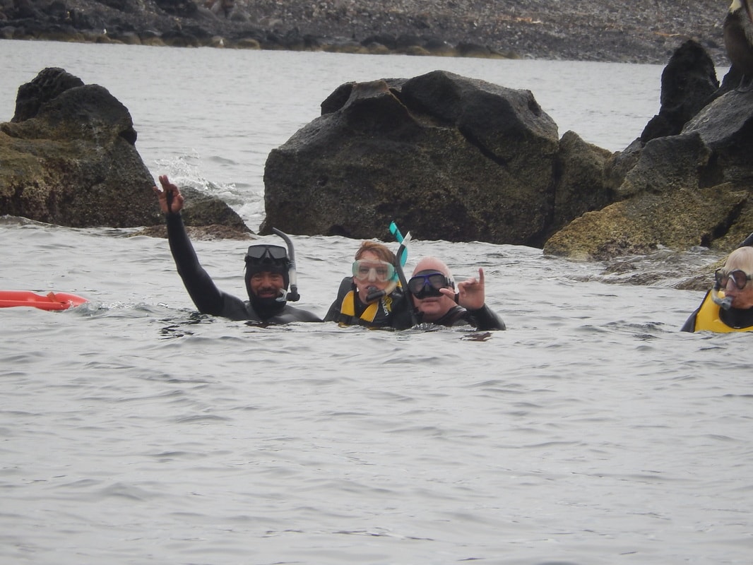 Guide and two guests in wetsuits waving and flashing shaka signs while snorkeling near rocky shores in the Gulf of California