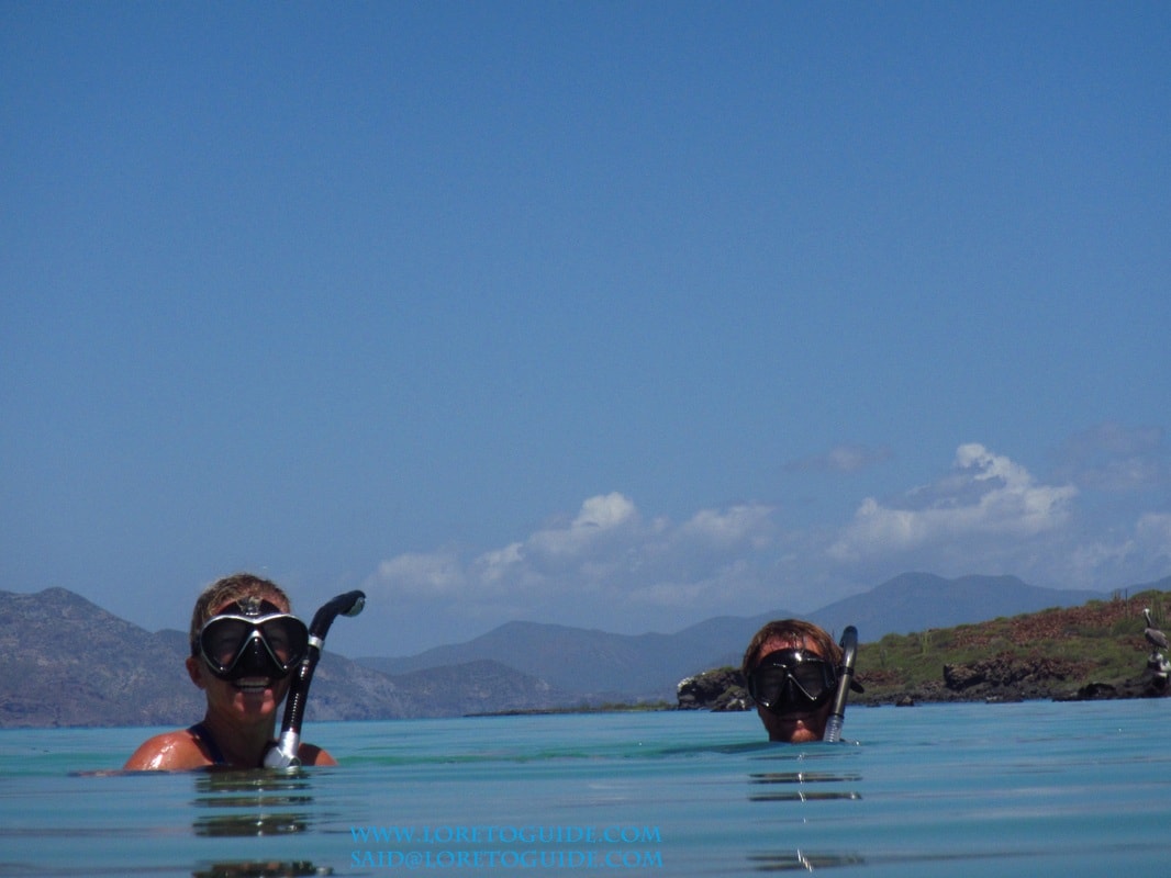 Two smiling snorkelers surfacing in crystal-clear water with islands and mountains behind them near Loreto