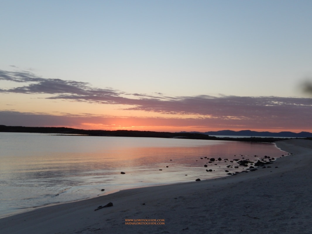 Glowing pink and orange sunset reflected on calm water along a white sand beach near Loreto