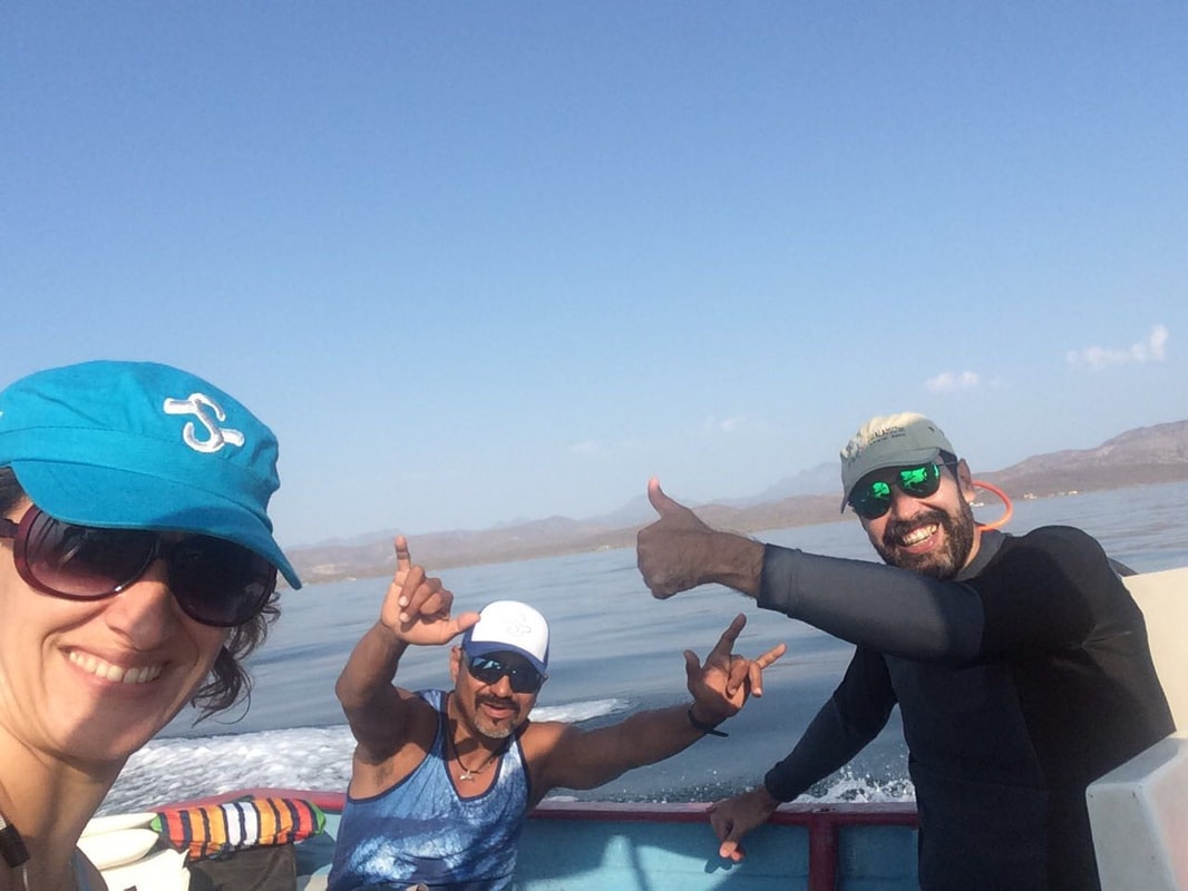 Three happy people giving thumbs up in a selfie on a panga with open Gulf of California water and distant sierra mountains behind them