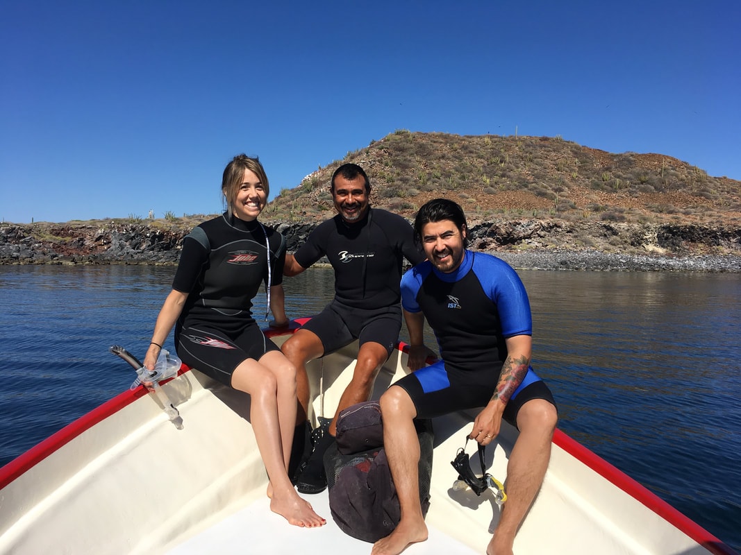 Three people in wetsuits sitting on a panga bow with a cactus-covered volcanic hill and calm blue water behind them near Loreto