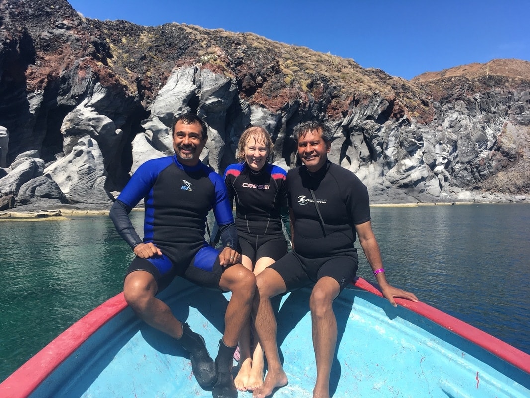 Three people in wetsuits sitting on the bow of a panga with dramatic volcanic rock cliffs behind them on a Loreto snorkeling tour