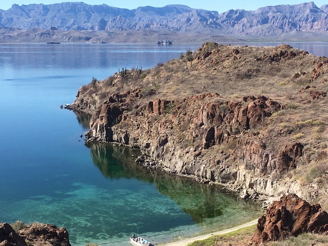 Aerial view of a rugged volcanic rocky peninsula with emerald and turquoise water, cactus, and the Sierra de la Giganta reflected behind near Loreto
