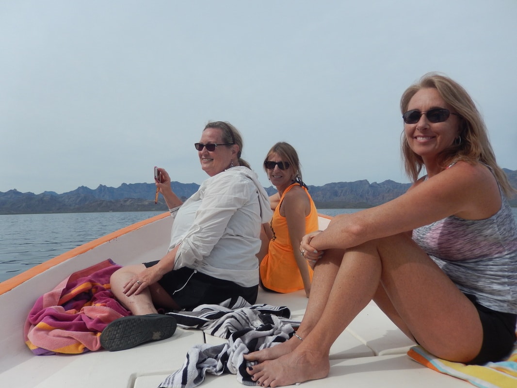 Six women laughing on a panga boat in front of a dramatic natural rock arch on a Loreto island tour