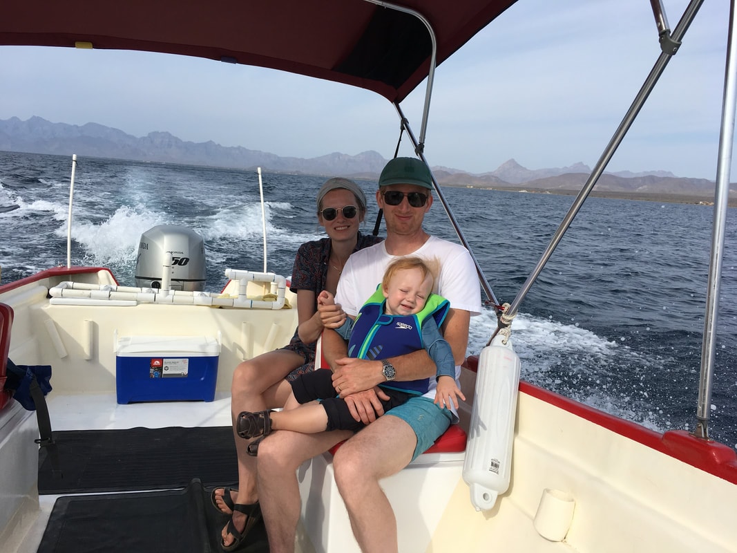 Young couple with toddler in life jacket smiling on a red-trimmed panga with sierra mountains rising behind them on a Loreto island tour